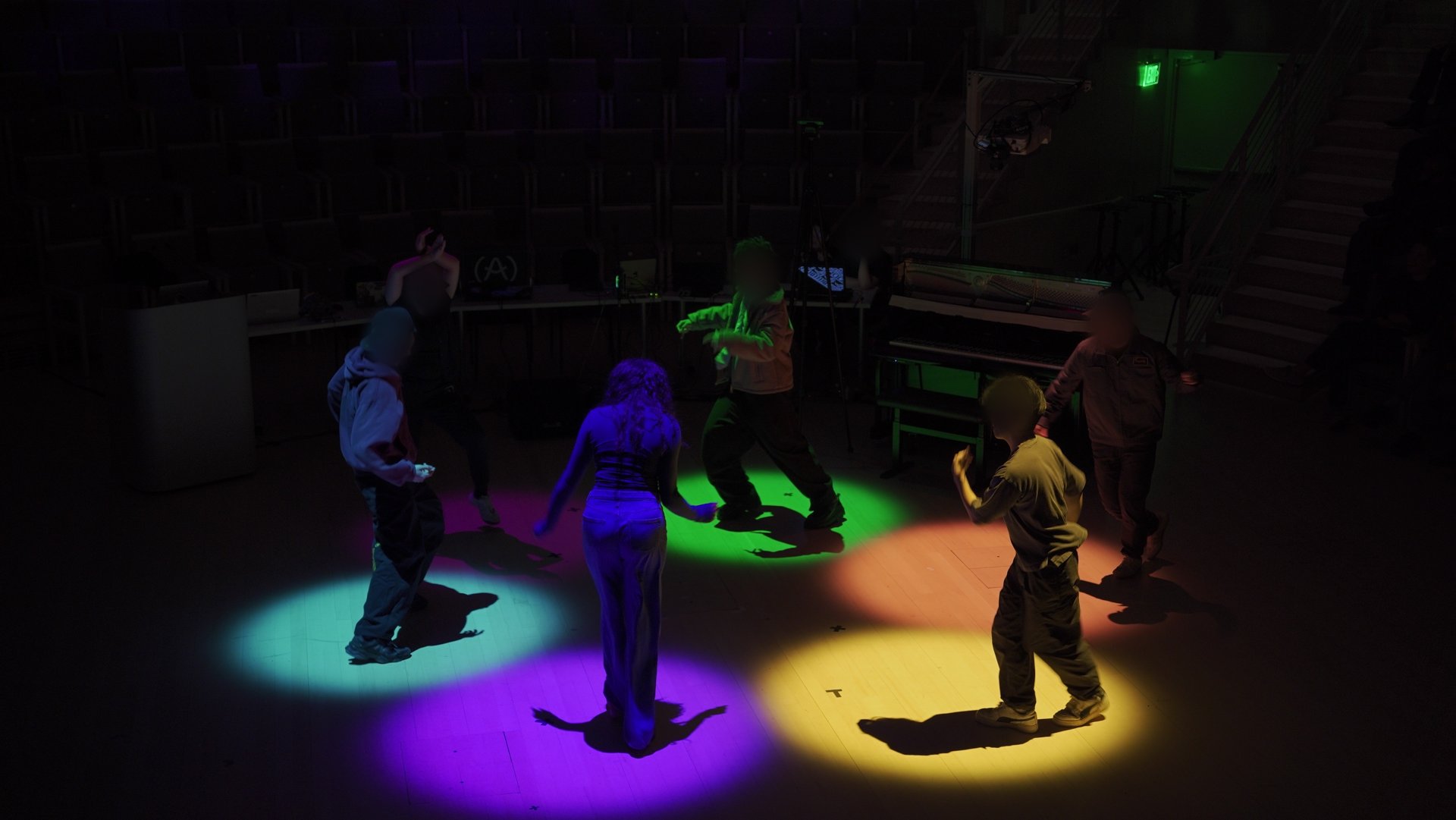 Six dancers in a dark concert hall, each standing in a differently colored pool of light on the floor; faces blurred.